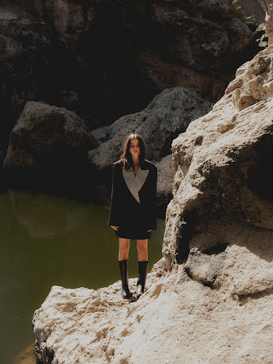 Woman in stylish blazer dress and boots poses on light rocks by green water in an outdoor canyon-like environment on a sunny day.
