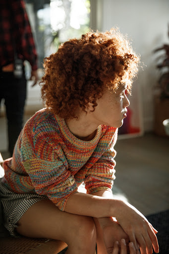 Woman with curly auburn hair sits indoors wearing a colorful patterned sweater and striped shorts, looking away from the camera.
