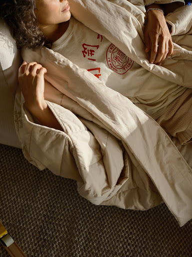 A young woman rests in a beige, quilted blanket indoors. The model is wearing a graphic t-shirt. Natural light enhances the cozy, relaxed setting.