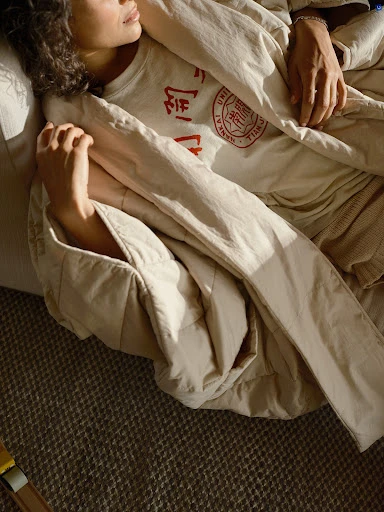 A young woman rests in a beige, quilted blanket indoors. The model is wearing a graphic t-shirt. Natural light enhances the cozy, relaxed setting.