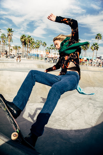 Male skateboarder performs a trick in an urban skatepark with palm trees and a sunny sky, wearing jeans and a patterned shirt. 