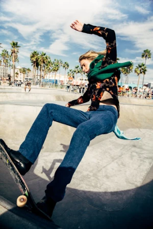 Male skateboarder performs a trick in an urban skatepark with palm trees and a sunny sky, wearing jeans and a patterned shirt. 