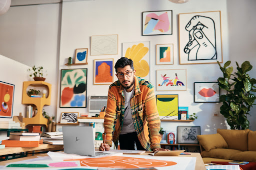 Man with glasses stands behind a desk with a laptop and drawings in an art studio, with colorful artwork hanging on the wall.