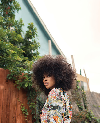 Portrait of a woman with large afro hair wearing a floral blazer, outdoors, in medium shot, posing and glancing back at the camera.