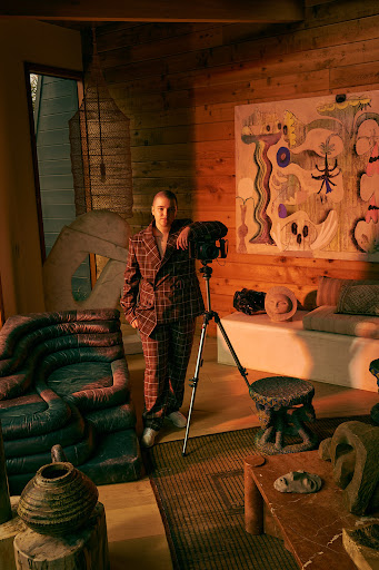 Adult female in plaid suit poses next to a camera on a tripod in a rustic wood paneled room with sculptures and abstract art.