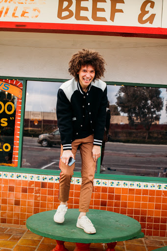 A young man stands on a green table outside a building, wearing a varsity jacket, brown pants, and white sneakers, smiling at the camera.