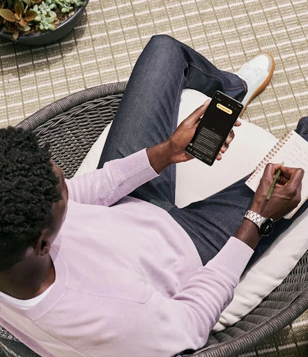 Overhead view of a man sitting outside using a smartphone and taking notes, dressed casually in jeans and a lavender sweater.