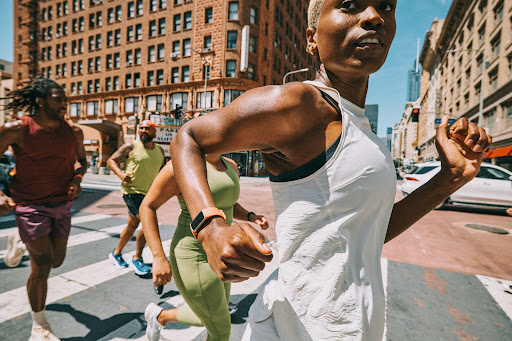 A diverse group of athletes, including an adult African-American woman in a white tank, runs across a city street on a sunny day.