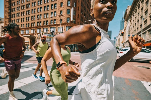 A diverse group of athletes, including an adult African-American woman in a white tank, runs across a city street on a sunny day.