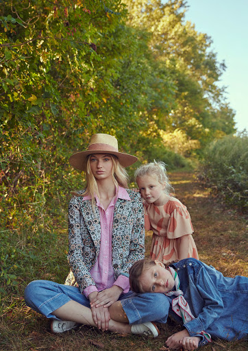 Woman and two children pose outdoors in a wooded area. The adult female wears a hat and patterned blazer. Two young girls in outdoor setting.