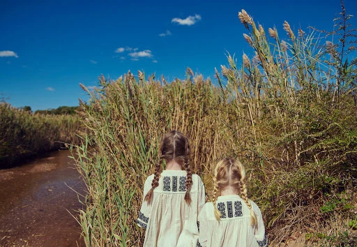 Two young girls with braids stand by a stream surrounded by tall grass under a clear blue sky. The girls wear matching embroidered tops.