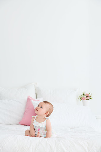 Infant sits on a white bed with a white wall in the background and looks up. There is a pink pillow and a bouquet of flowers.