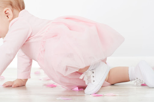 Baby girl in pink dress crawls on wooden floor with rose petals, showcasing her white sneakers and socks against a white background.