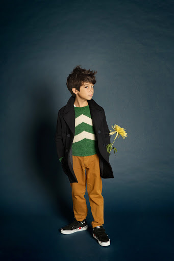 Full shot of a child dressed in a green and white chevron sweater, brown pants, and a dark jacket, holding a flower against a dark blue backdrop.