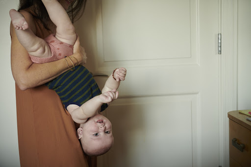 Adult is holding a baby upside down indoors, wearing a dress and the baby has a striped shirt. The background is a white door.