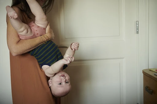 Adult is holding a baby upside down indoors, wearing a dress and the baby has a striped shirt. The background is a white door.