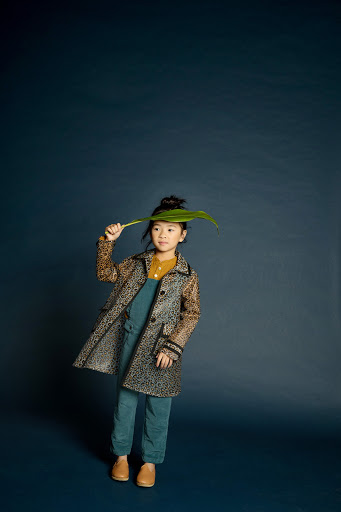 Young girl in an animal-print coat stands on a dark blue background, holding a large green leaf over her head in a studio portrait.