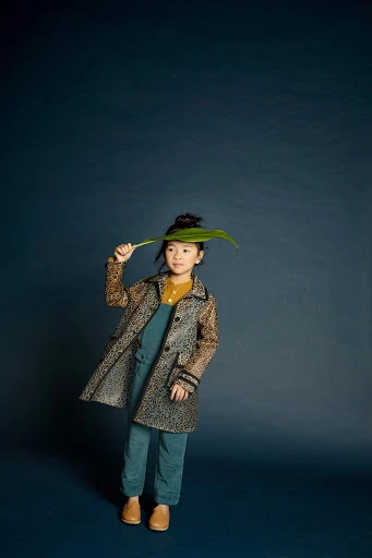 Young girl in an animal-print coat stands on a dark blue background, holding a large green leaf over her head in a studio portrait.