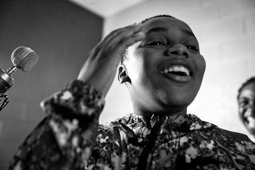 Monochrome shot of a young Black woman singing into a microphone, hand raised, with a second woman smiling softly in the background.