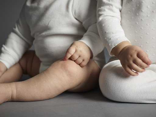 Close-up studio shot of two babies wearing white bodysuits, focusing on their hands and knees against a neutral grey background.