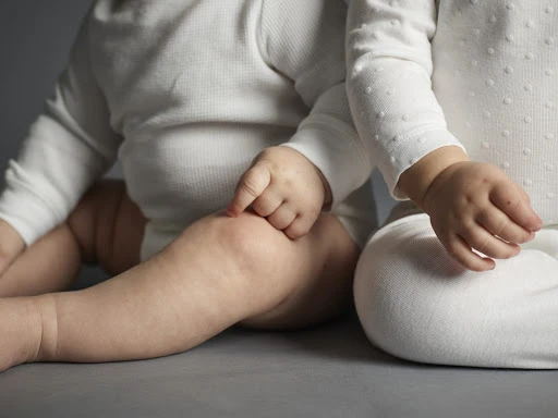 Close-up studio shot of two babies wearing white bodysuits, focusing on their hands and knees against a neutral grey background.