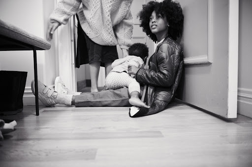 Black and white photo of a child sitting with an adult woman as another adult walks by in a doorway inside a home.