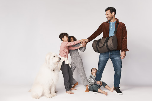 Father in a brown corduroy jacket holding a duffel bag, surrounded by three children and a large white dog