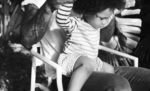 Black-and-white photo of a child in striped clothing sitting on an adult's lap, with lush foliage in the blurred background.