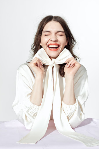 A woman smiles brightly, wearing a white top and large bow. She is indoors with a white background and a lavender table surface.