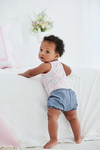 Adorable African American baby girl in a floral print top and denim bloomer shorts, standing and holding onto a white textured bed.