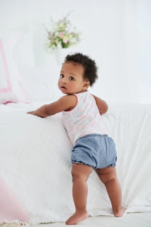 Adorable African American baby girl in a floral print top and denim bloomer shorts, standing and holding onto a white textured bed.