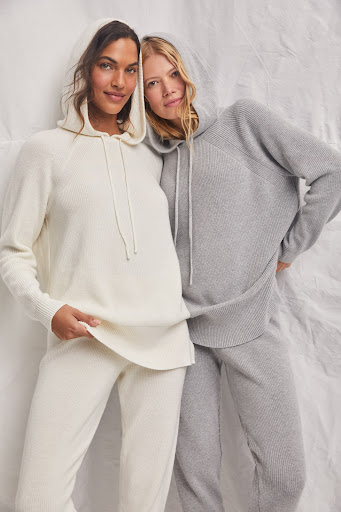 Two women pose in matching hooded sweaters and pants in a studio setting, showcasing comfortable and stylish loungewear.