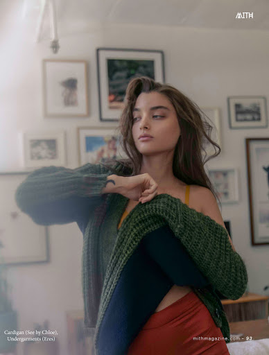 Portrait of a woman wearing a green cardigan, orange shorts, and black tights, sitting indoors in front of art-covered walls.