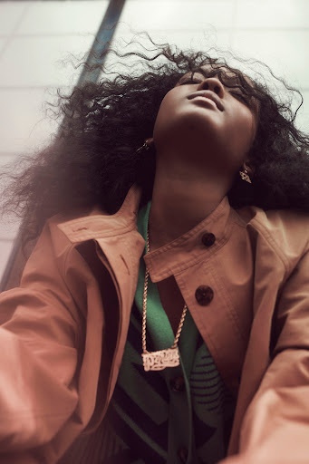 Adult woman with curly black hair and a gold necklace, wearing a trench coat and a green top, looking up in a close-up shot.