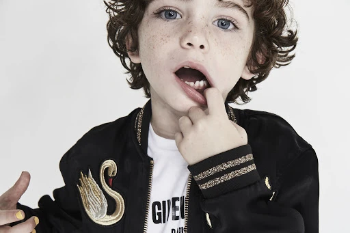Close-up portrait of a child with blue eyes and freckles wearing a black embroidered bomber jacket against a white background.