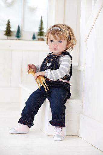 A young child with blonde hair is sitting on white stairs indoors, wearing denim overalls, a striped shirt, and pink sneakers, holding a deer figurine.