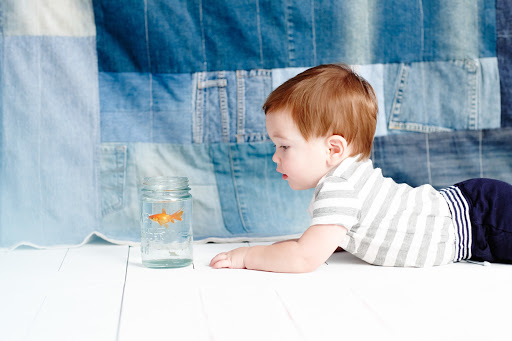Baby boy lying on a white floor looking at a goldfish in a clear glass jar, with denim patchwork background, showcasing childhood wonder and curiosity.