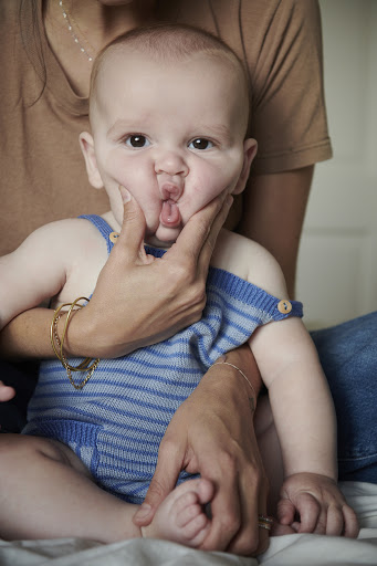 Adorable baby with funny face held by adult indoors, wearing blue striped romper and gold jewelry, mother and child portrait.