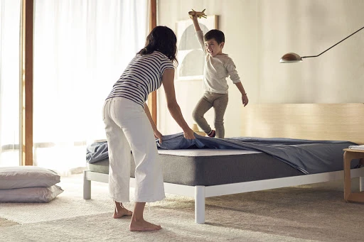 Woman makes a bed indoors in a sunlit bedroom as a young child plays with a toy airplane, with a light rug on the floor.