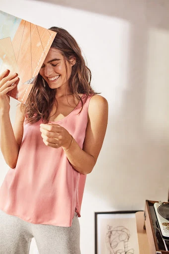 A cheerful young woman in a pink top stands indoors, playfully holding a vinyl record over her face while smiling brightly.