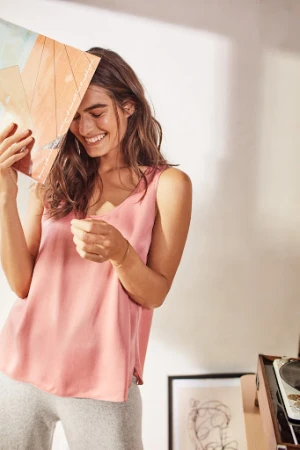 A cheerful young woman in a pink top stands indoors, playfully holding a vinyl record over her face while smiling brightly.