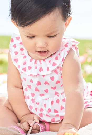 Adorable baby girl sits outdoors on a blanket wearing a white romper with pink hearts, looking down, with green grass in the background.