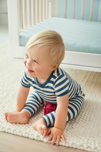 A smiling blond toddler wearing a striped pajama set sits on a white rug indoors near a baby crib in a cozy and cheerful setting.