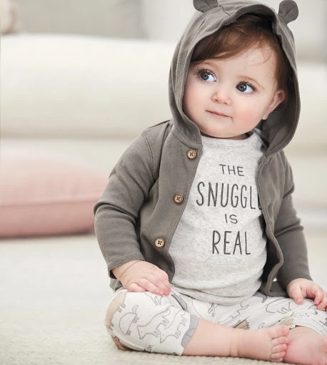 Cute baby with big eyes wears a hooded jacket and printed leggings, sitting indoors on a light carpet with neutral colors, looking off-camera.
