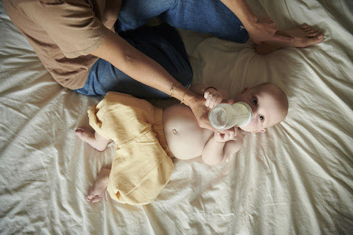 Infant in diaper shorts being fed a bottle by an adult, lying on a white sheet. Gentle natural light, close-up shot, high angle.