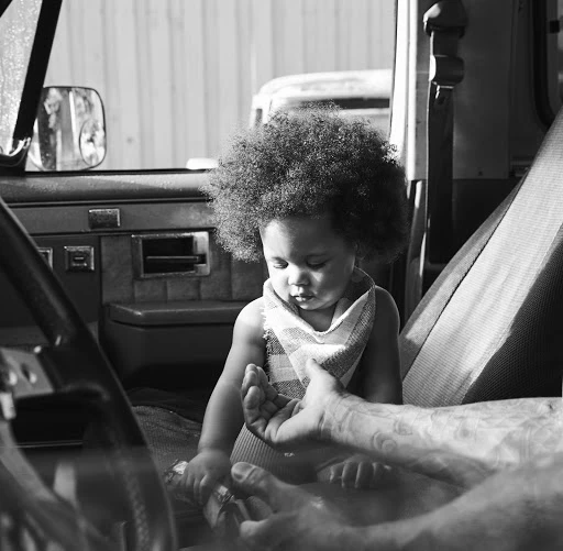 Black and white portrait of a baby sitting inside a vehicle with an adult's hands visible, creating a tender, intimate scene.