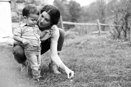 Black and white portrait of a woman crouching outdoors, interacting with a young child standing in grass.