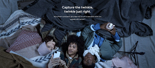 Three people laying on a bed with blankets. One is holding a phone, another is holding a cup. Overhead shot with warm, cozy lighting.
