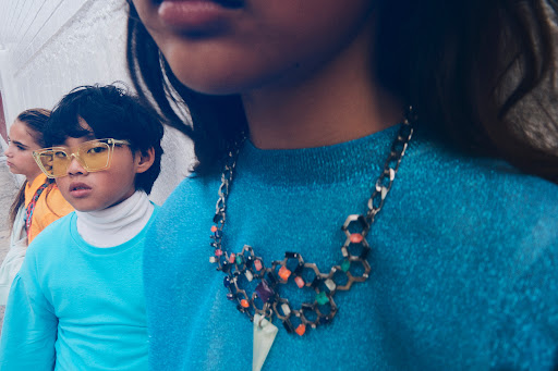 A group of three children are wearing colorful clothing and fashionable accessories, standing outdoors with a neutral background.