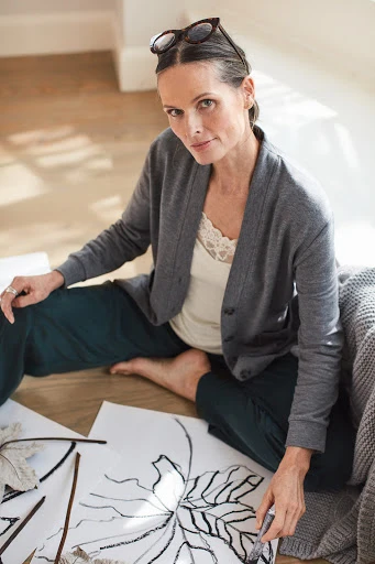 Mature woman sits barefoot on the floor drawing botanical line art with black marker. She wears a grey cardigan and dark pants.